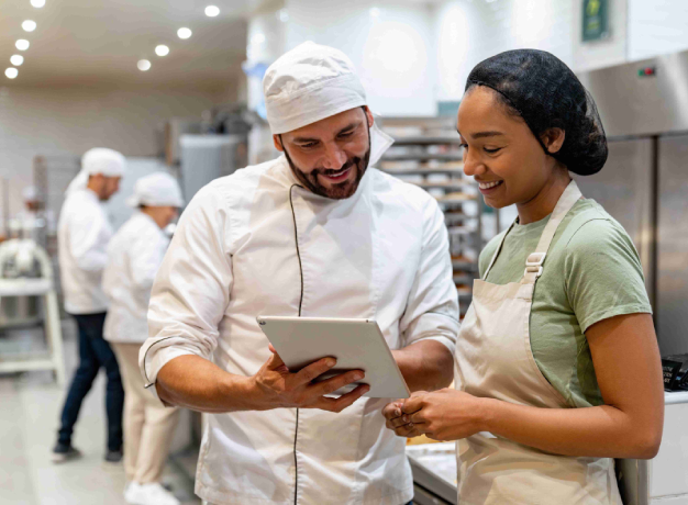 Bakery workers reviewing information on a tablet in a commercial kitchen, highlighting teamwork, food production, and workplace collaboration.